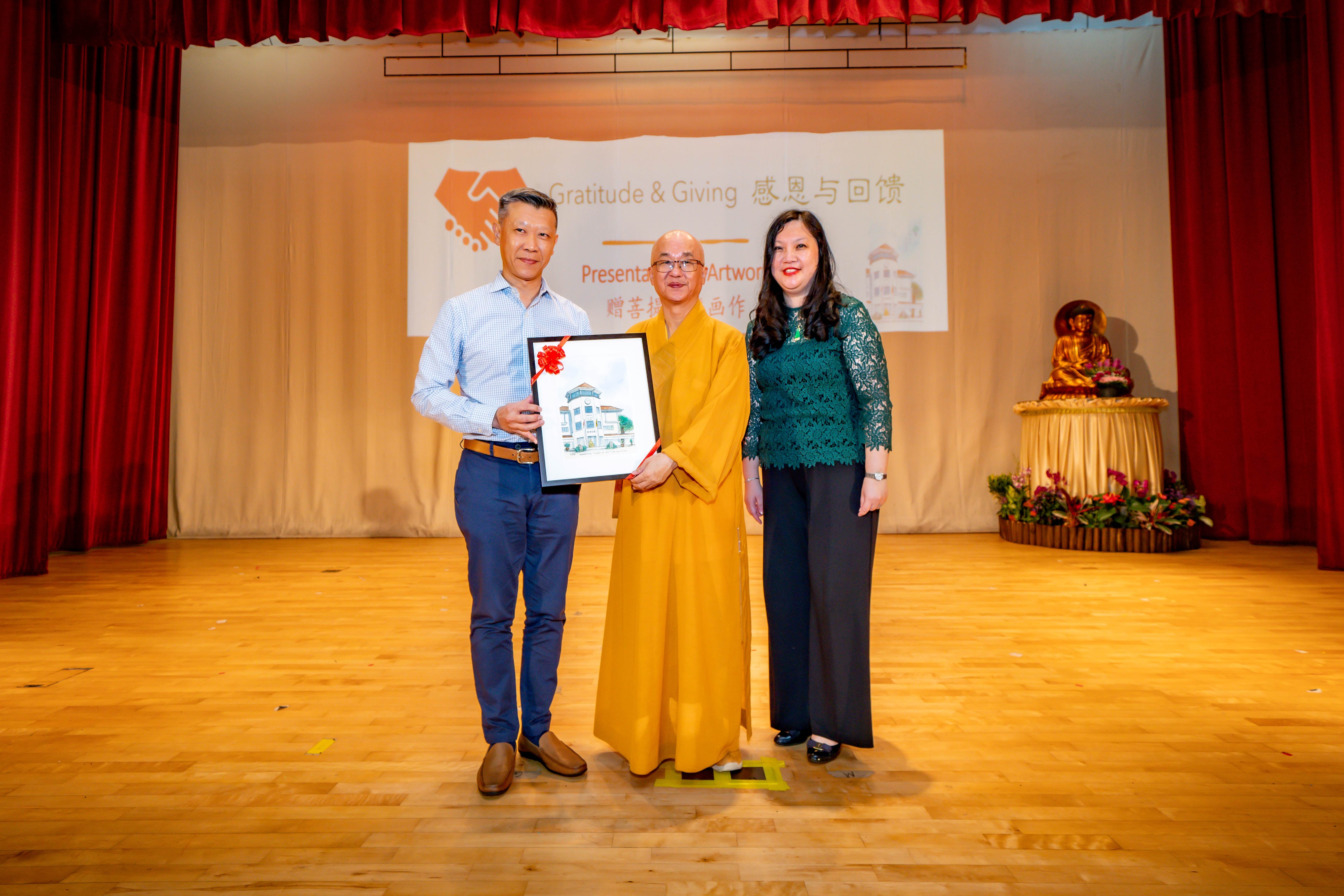 Three people on a stage pose with a framed artwork at a Gratitude and Giving event, with a Buddhist monk in yellow robes centered.
